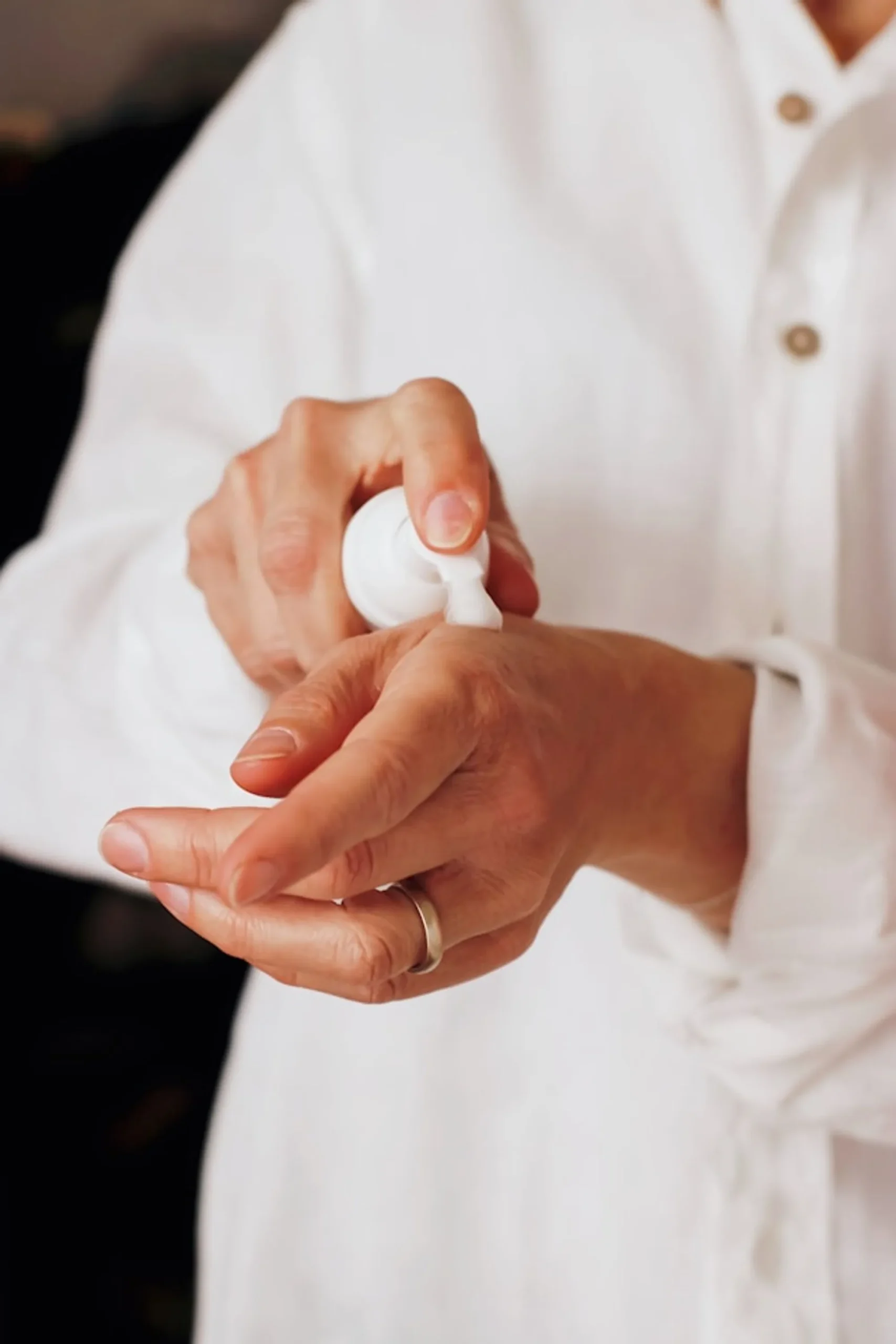   a woman with a white shirt, and a gold ring on her finger, putting a skincare product on the palm.