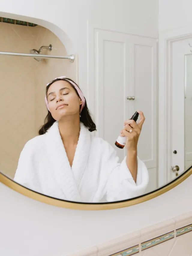 a reflection of a lady with in a white bathrobe and a pink head band, behind a white door, spraying skin care product on her face on a mirror.