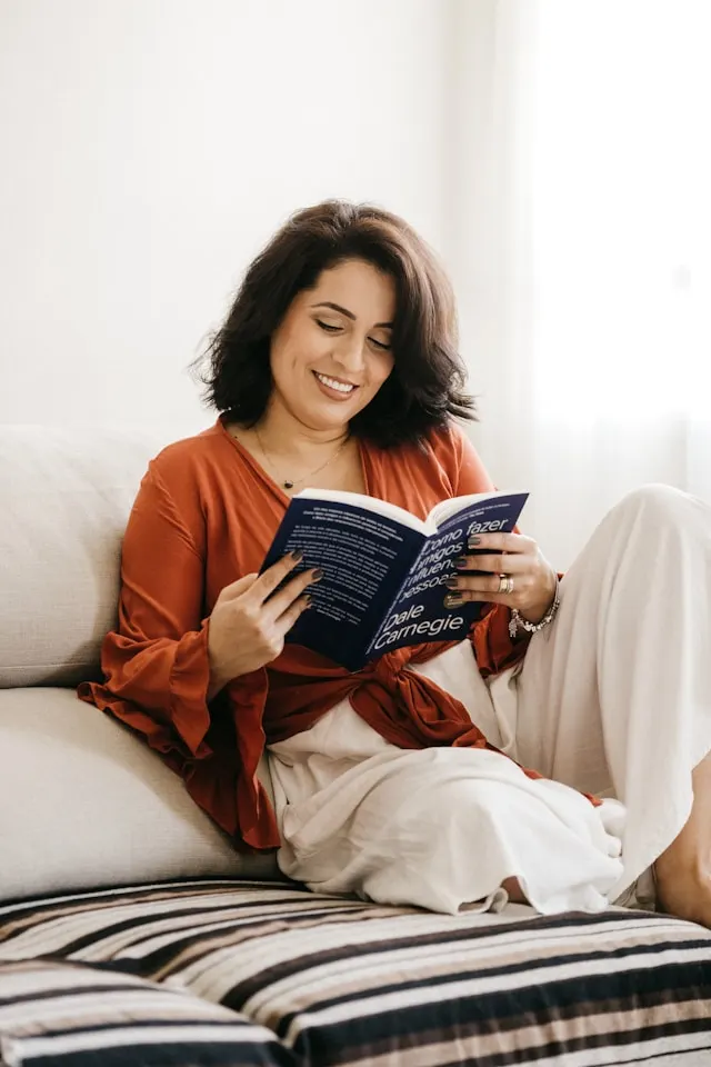 Alt text: A lady with a brown long sleeve top and white palazzo pants, sitting on  white sofa with white and black strips, reading blue a book. 